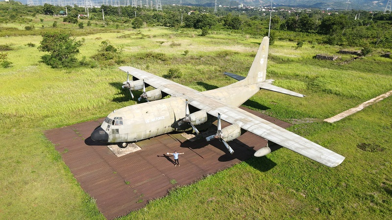 A four-engine propeller transport aircraft in faded grey paint, parked on a square of rusted steel matting in a grassy field, with a person standing beside it