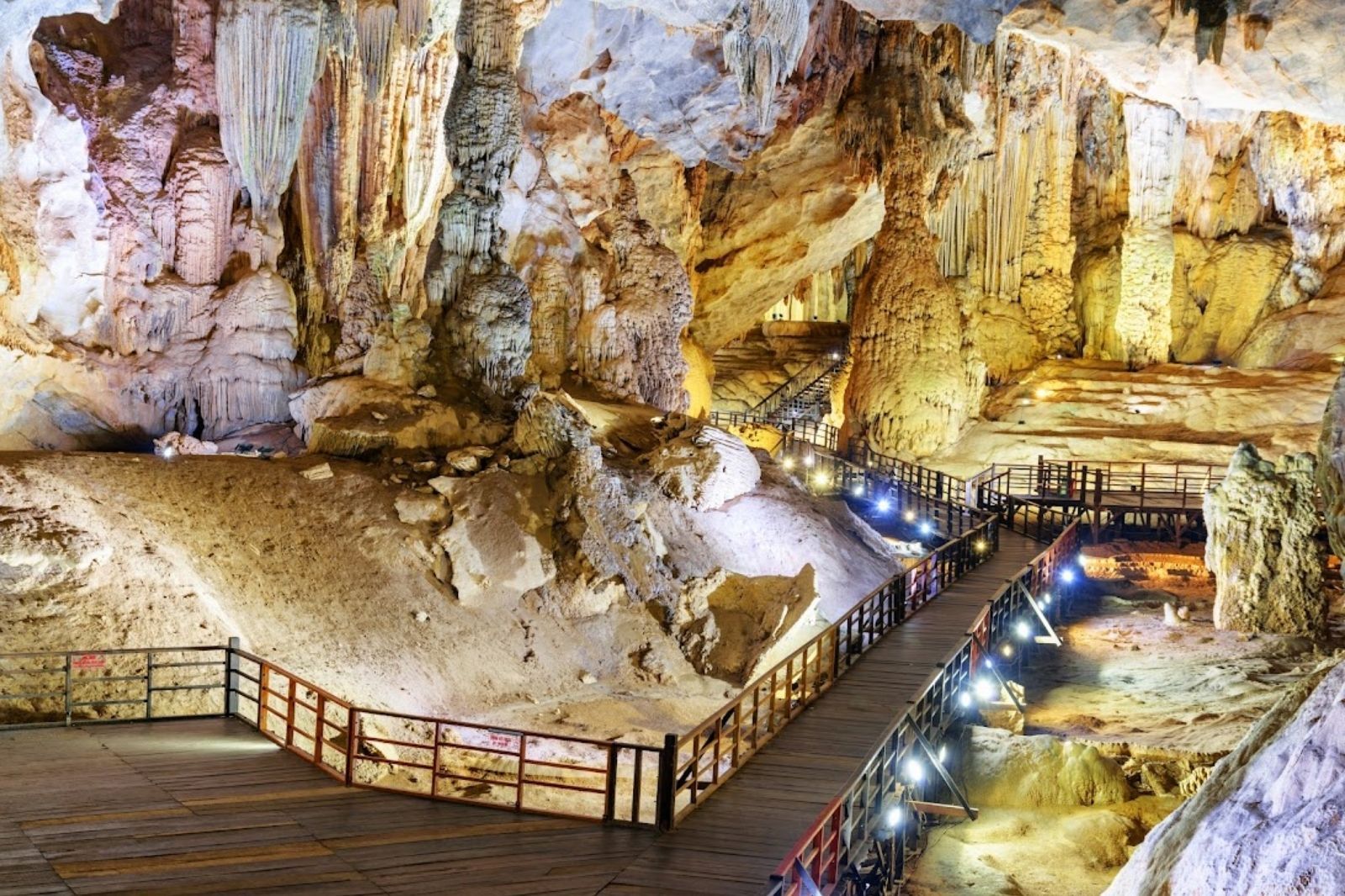 A lit wooden boardwalk with railings winding between huge pale stalactites and stalagmites inside a cave