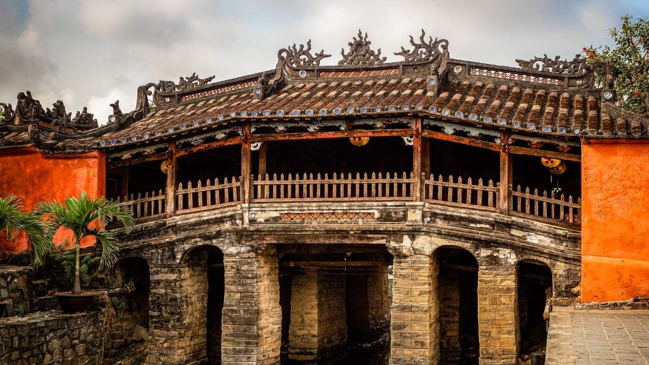 A covered wooden bridge with a curved tiled roof and carved roof ornaments, standing on stone arches between orange-walled buildings