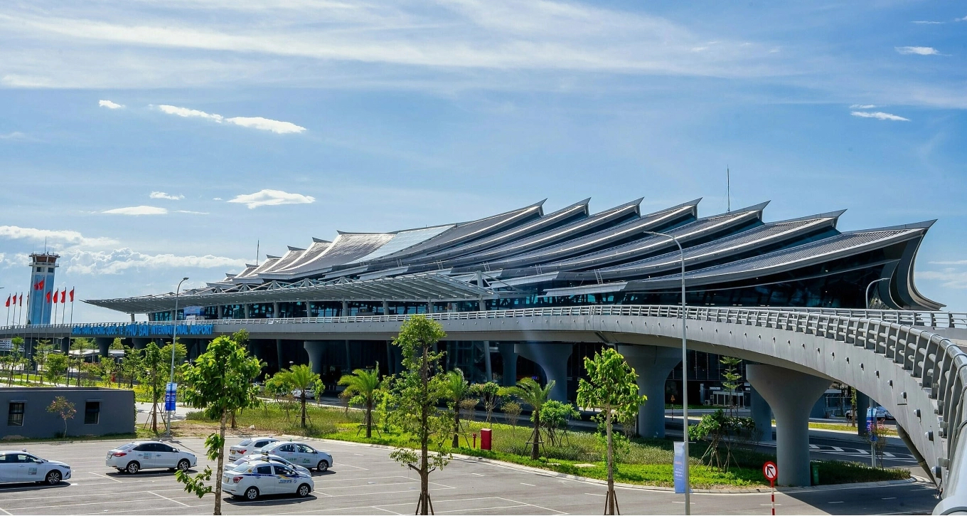 A modern airport terminal with a roof built as a row of curved, upswept fins, an elevated approach road, a control tower, young palm trees and white taxis in the car park in front