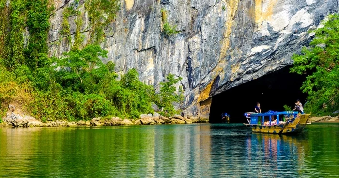A tourist boat on a still green river at the dark mouth of a cave in a tall limestone cliff