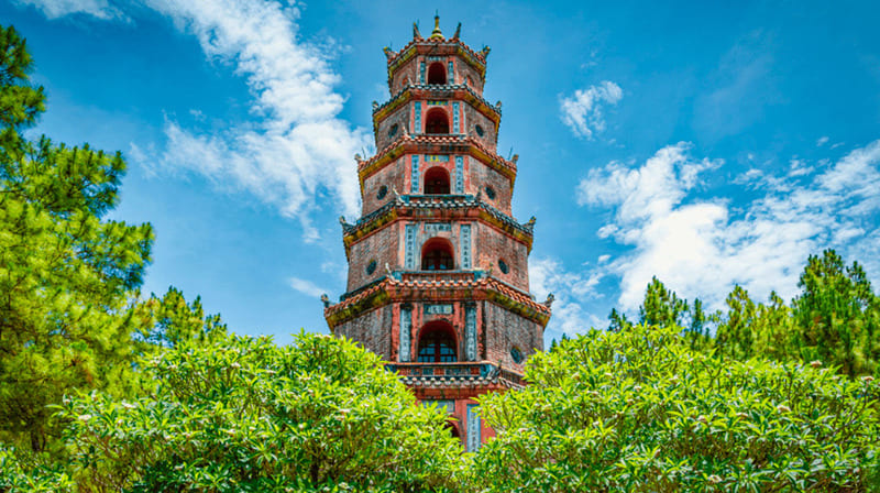 A seven-tiered brick tower with ornate tiled eaves rising above flowering trees and pines under a blue sky