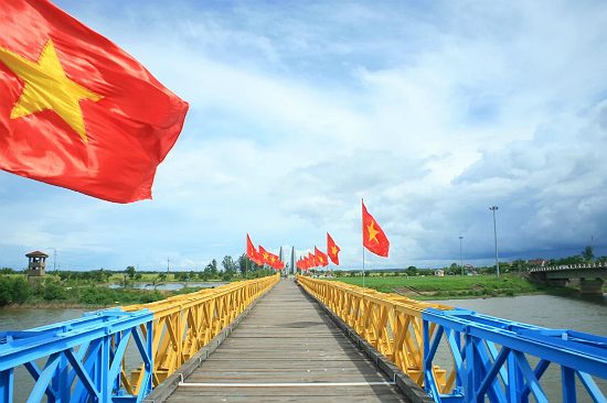 A bridge with a wooden deck and blue and yellow steel railings, lined with red flags bearing a yellow star, crossing a wide river under a cloudy sky