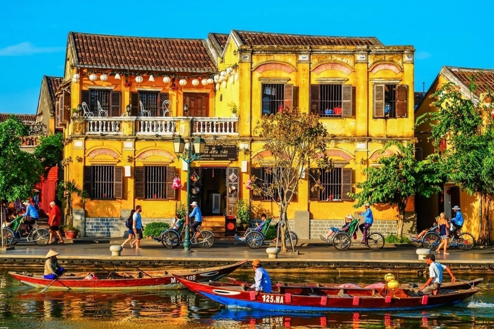 Two-storey yellow shophouses with brown tiled roofs and strings of white lanterns along a riverside walkway, with cyclos and pedestrians in front and wooden boats on the river