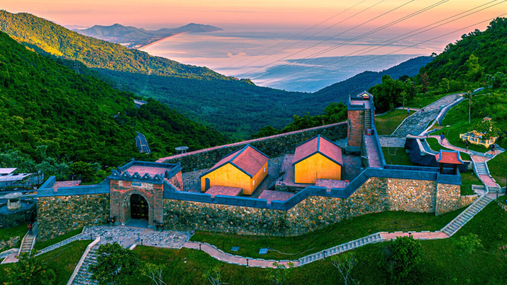 A stone fort with a brick gatehouse and two yellow buildings with red roofs on a green mountainside, a winding road below and a long beach and bay in the distance under a pink and orange sky