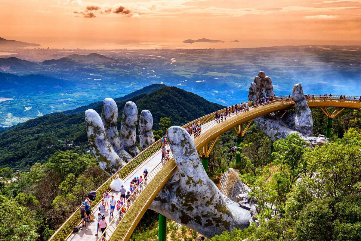 A curved pedestrian bridge with golden railings held up by two giant stone hands, crowded with visitors, on a forested mountainside above a wide plain and the sea