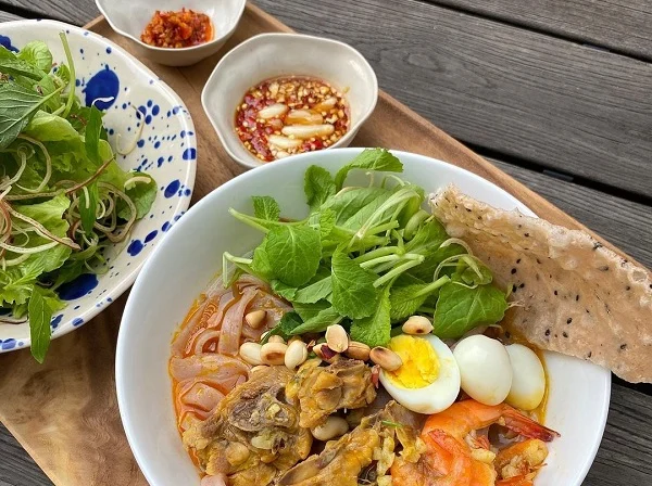 A bowl of flat noodles in a little orange broth, topped with prawns, quail eggs, peanuts, meat and fresh greens, with a sesame rice cracker, a plate of herbs and two small dishes of chilli on a wooden tray