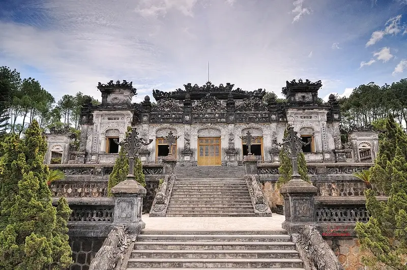 A grey stone mausoleum with carved pillars and ornate rooflines at the top of several long flights of stone steps, flanked by clipped cypress trees