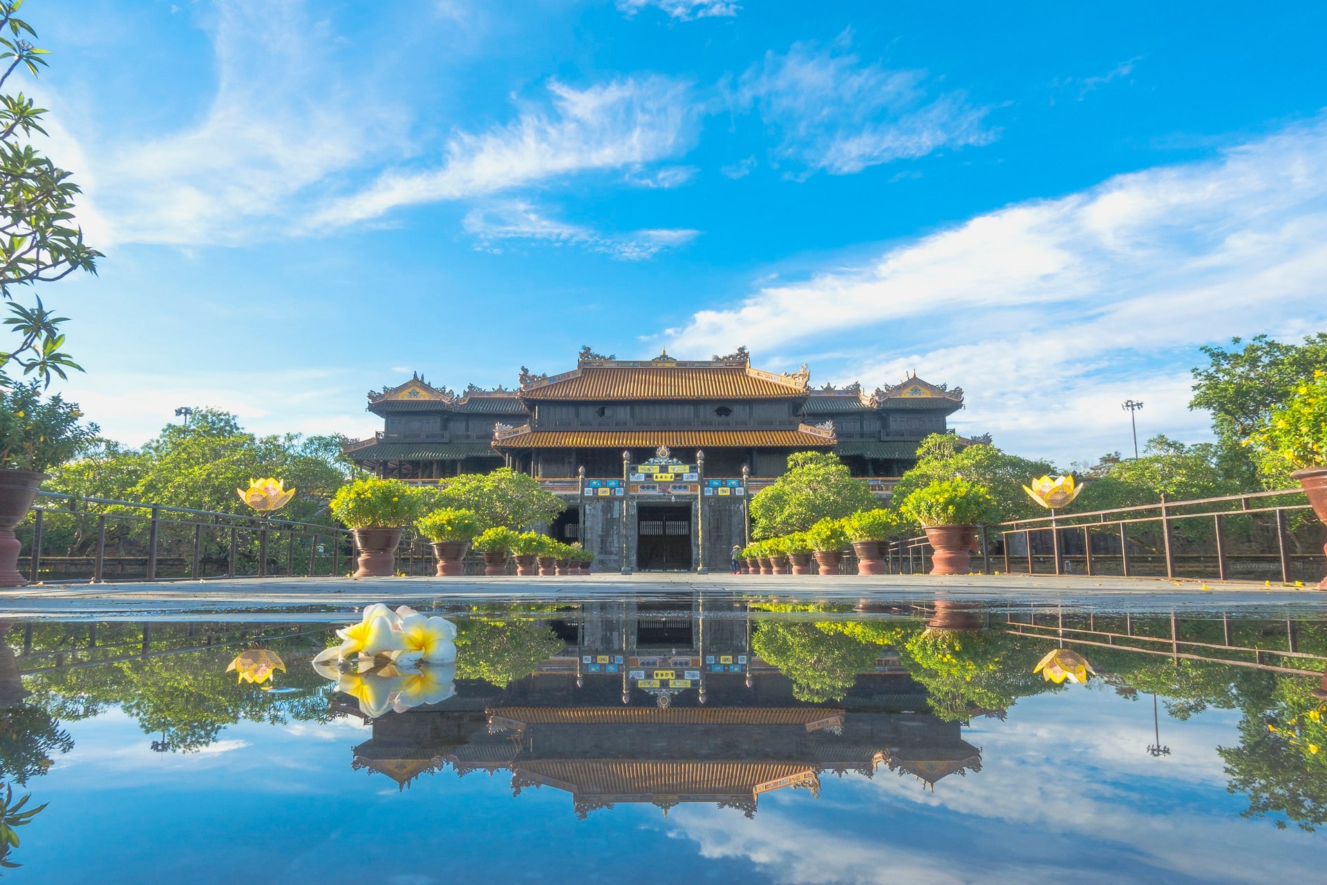 A multi-roofed wooden gatehouse with golden-tiled roofs above a stone base, flanked by potted trees, reflected in a pool of water with a frangipani flower in the foreground