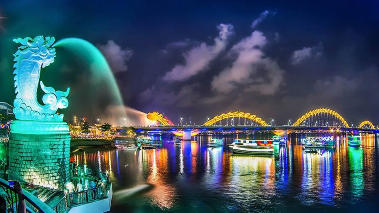 A river at night with a bridge of illuminated yellow arches, brightly lit tour boats and their reflections, and a lit statue of a fish-dragon spouting water in the foreground