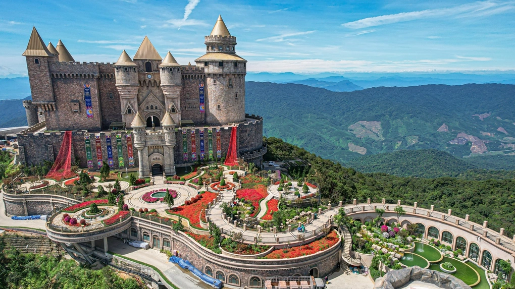 A mock-medieval stone castle with pointed towers above terraced flower gardens, on a mountaintop surrounded by forested hills